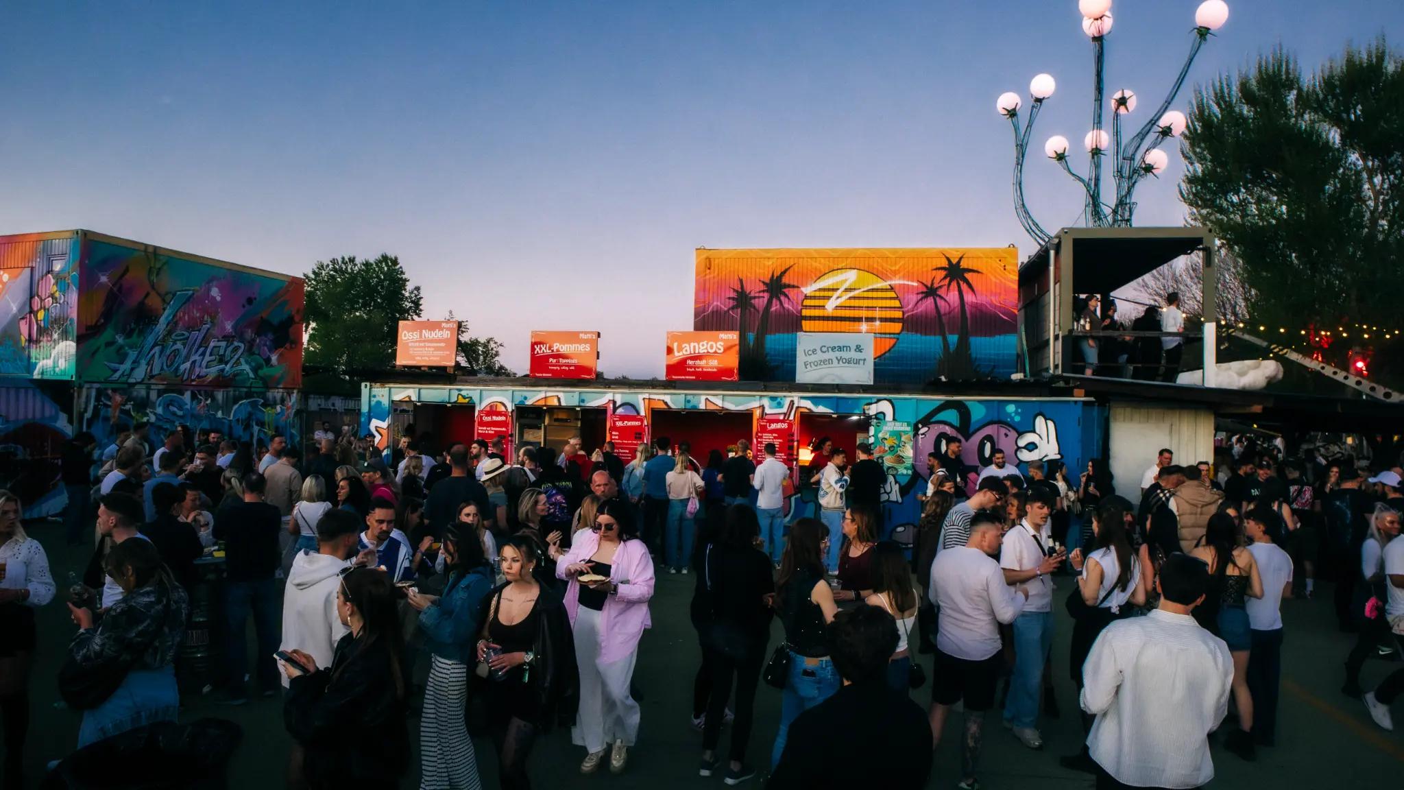 Belebter Food Court mit graffitibemalten Verkaufsständen, Lichterdekoration und vielen Personen
